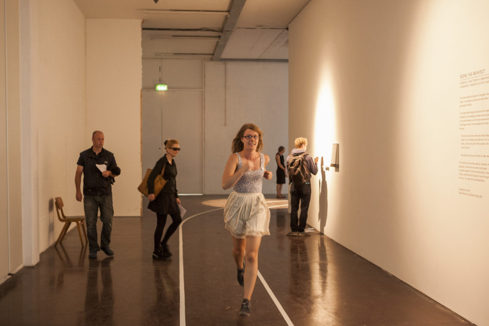 A white walled gallery corridor is marked out with a running track around which a girl is running, past visitors who are exploring the exhibition