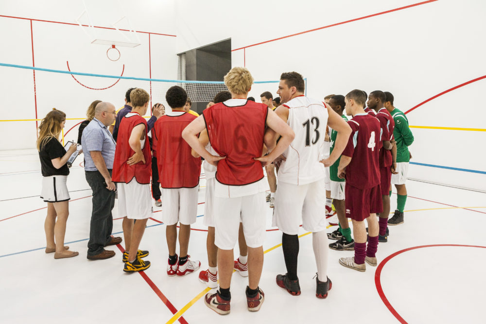 A large group of people wearing sports kit and coloured tabards are gathered together in a white walled gallery marked out with colourful lines like a sports hall