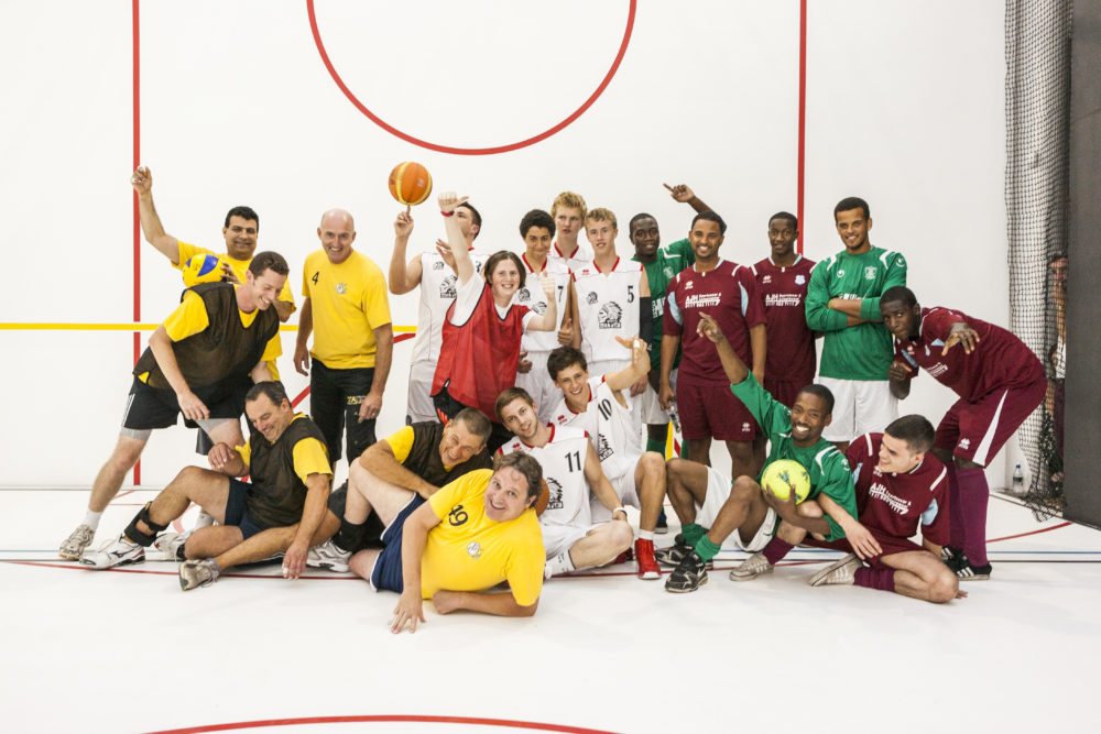 A large group of people wearing sports kit and coloured tabards are gathered together for a photograph in a white walled gallery marked out with colourful lines like a sports hall