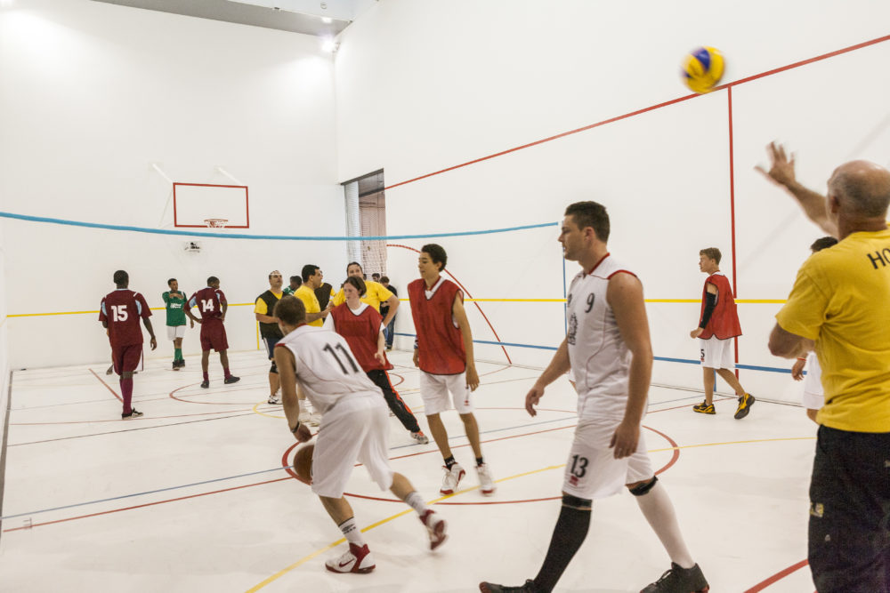 A sports team of people wearing sports kit for playing basketball are gathered together in a white walled gallery marked out with colourful lines like a sports hall