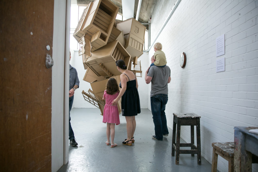 Visitors inside an artists' studio looking at an installation of wooden structures that look like abstracted houses tumbling from the ceiling.