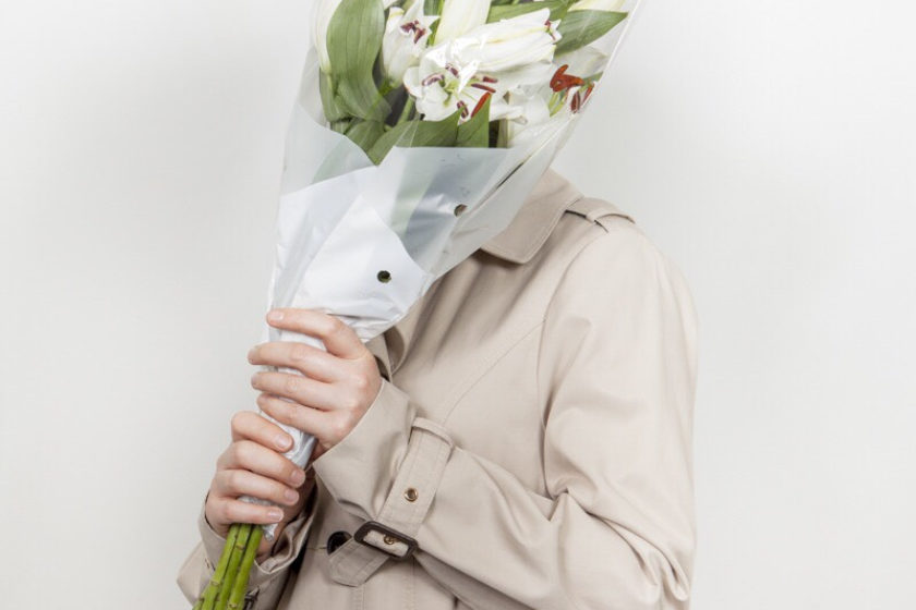 Photograph of a man in a beige mackintosh jacket holding a bunch of white lilies to hide his face