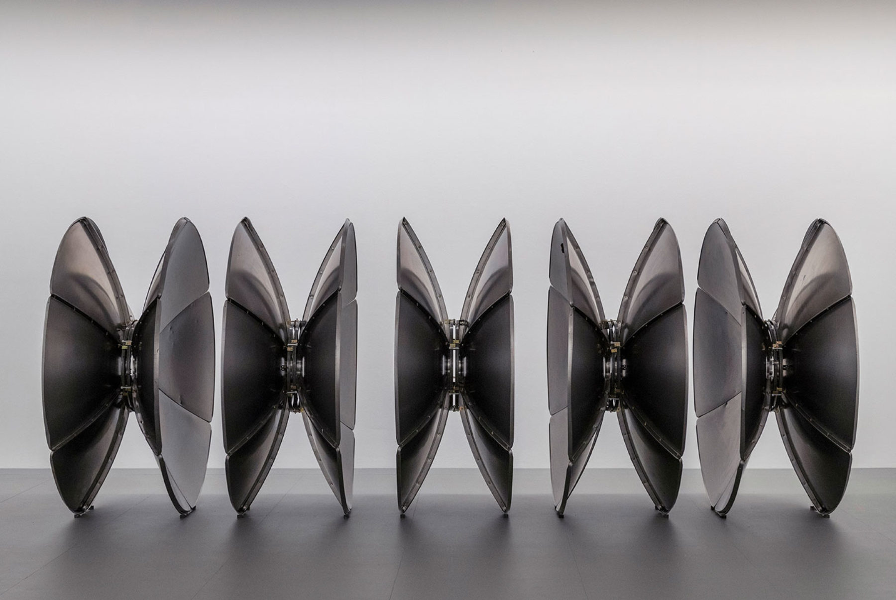 Photograph of five round metal sculptures on the floor in a gallery space, a white wall behind them.