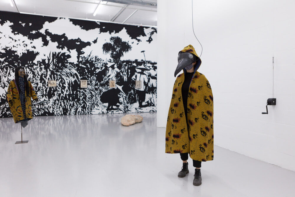 Gallery view. A figure on the right wears a bird-like VR mask and a patterned yellow fabric cloak. Behind this, a large black and white mural depicts Chinese agricultural workers.