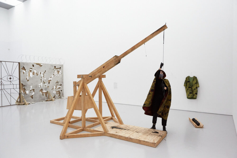 A figure wearing a mask and cloak approaches the trebuchet, a large wooden catapult structure.