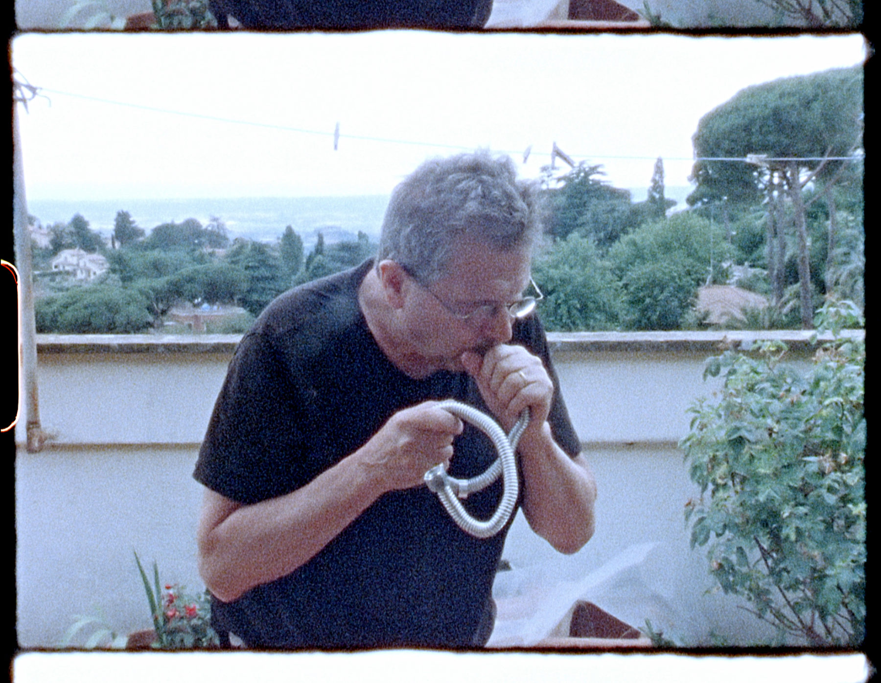 Film still featuring a man with white hair and round glasses and wearing a black tshirt, blowing into a flexible hose.