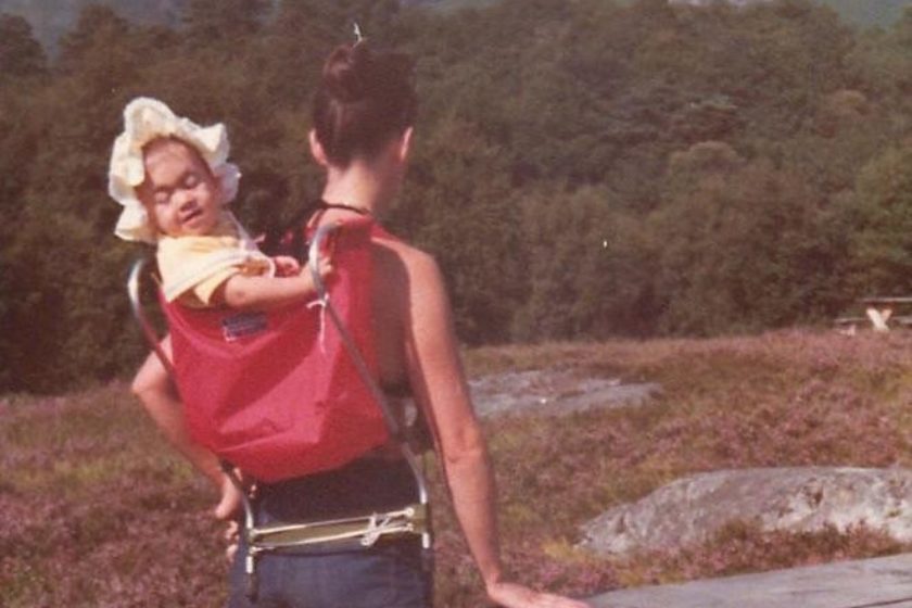 A photograph of a mother standing by a picnic bench, wearing shorts and a vest, with a child in a carrier on her back. The baby is wearing a large white bonnet.