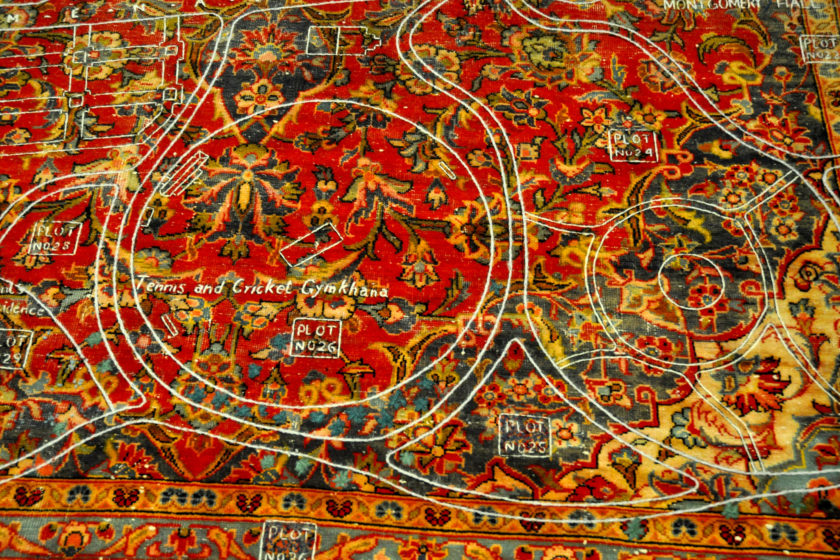 White thread stitched upon an antique Kashan carpet. The carpet has a flowery pattern with red, green, yellow, and blue colours. It is threadbare in some places. The white thread depicts a map with written annotations including 