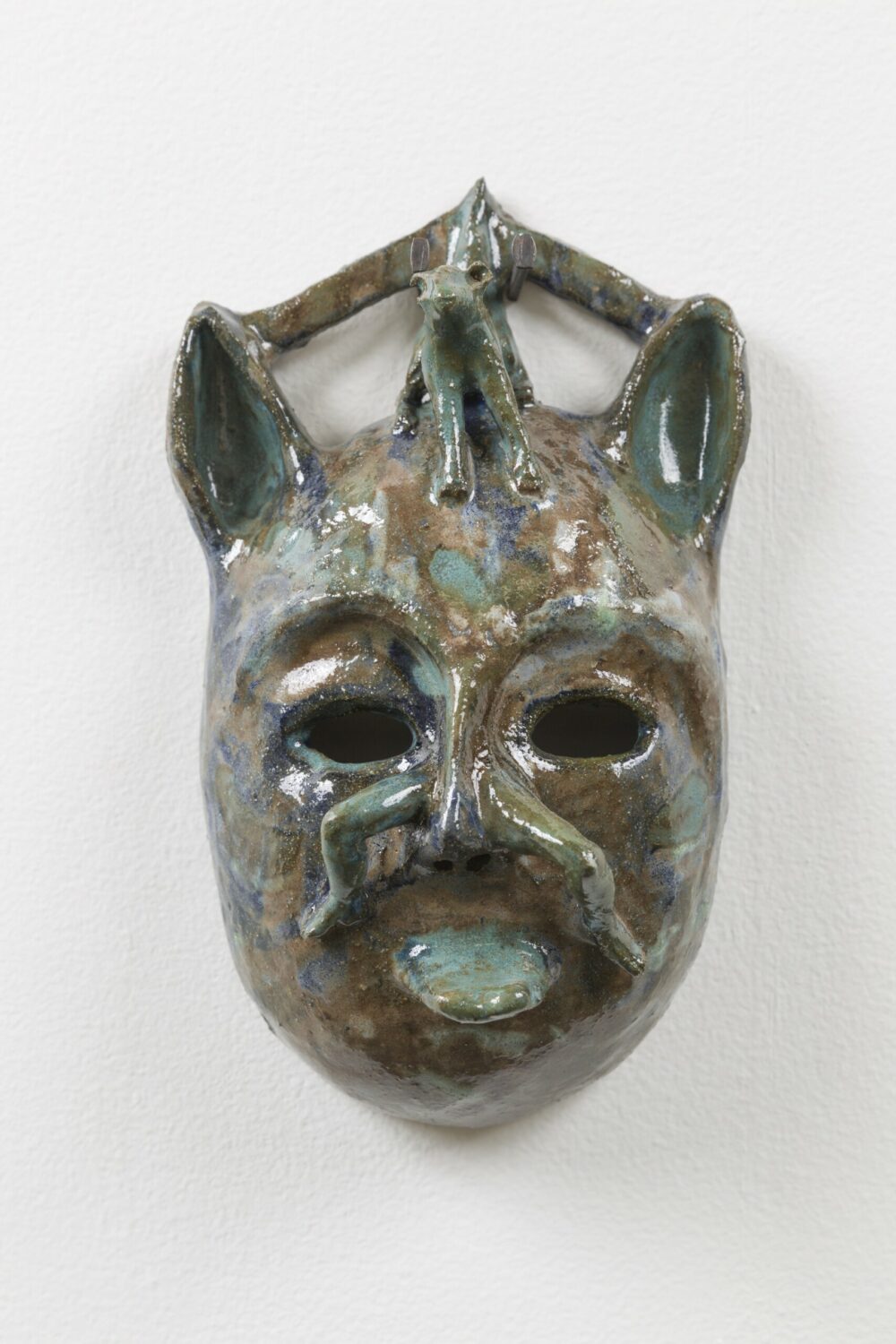 A glazed ceramic mask in silver and bronze tones, with ears and hollow eye sockets, and the centre of the face is shaped like the form of a small figure.