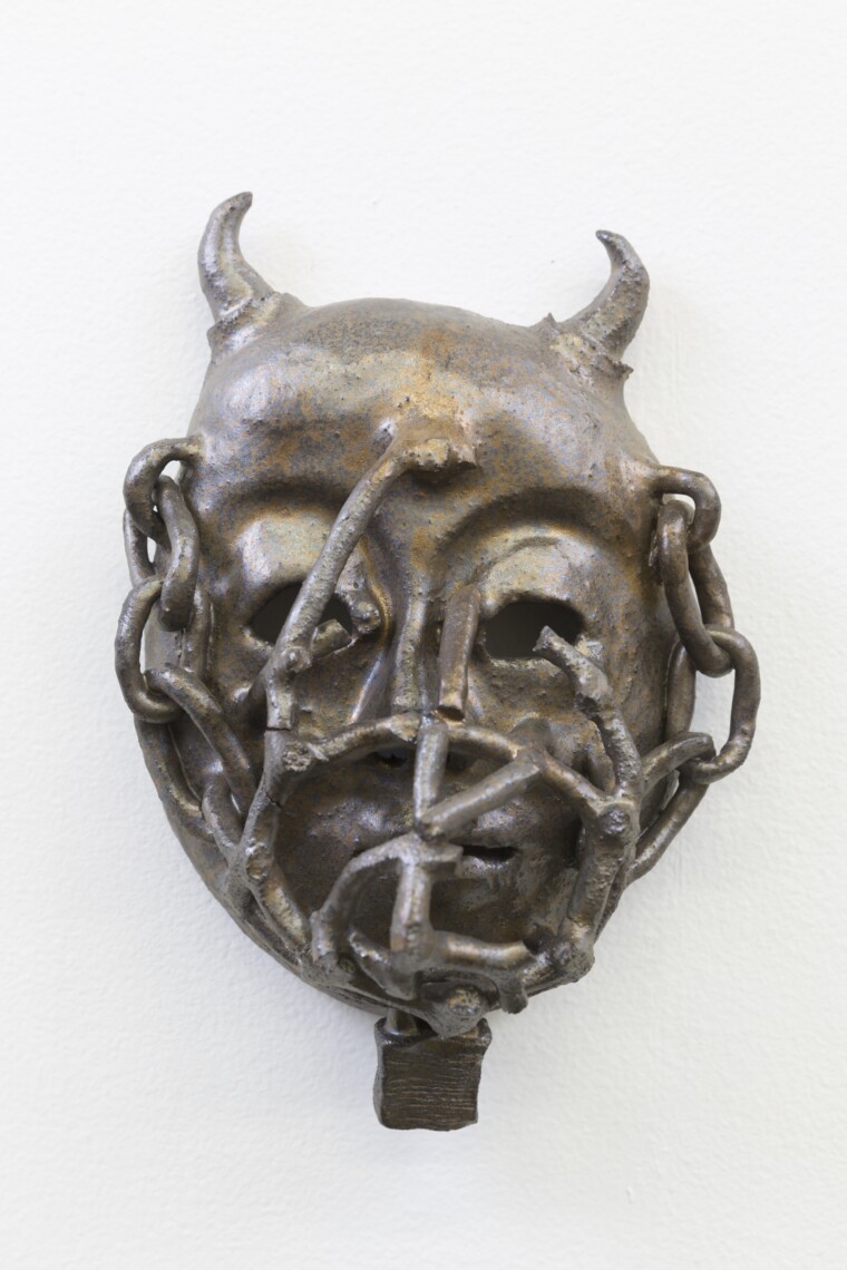 A glazed ceramic mask in silver and bronze tones, featuring a head with small horns, covered in chains which enter into the hollow eye sockets.