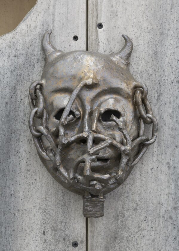 A glazed ceramic mask in silver and bronze tones, featuring a head with small horns, covered in chains which enter into the hollow eye sockets.