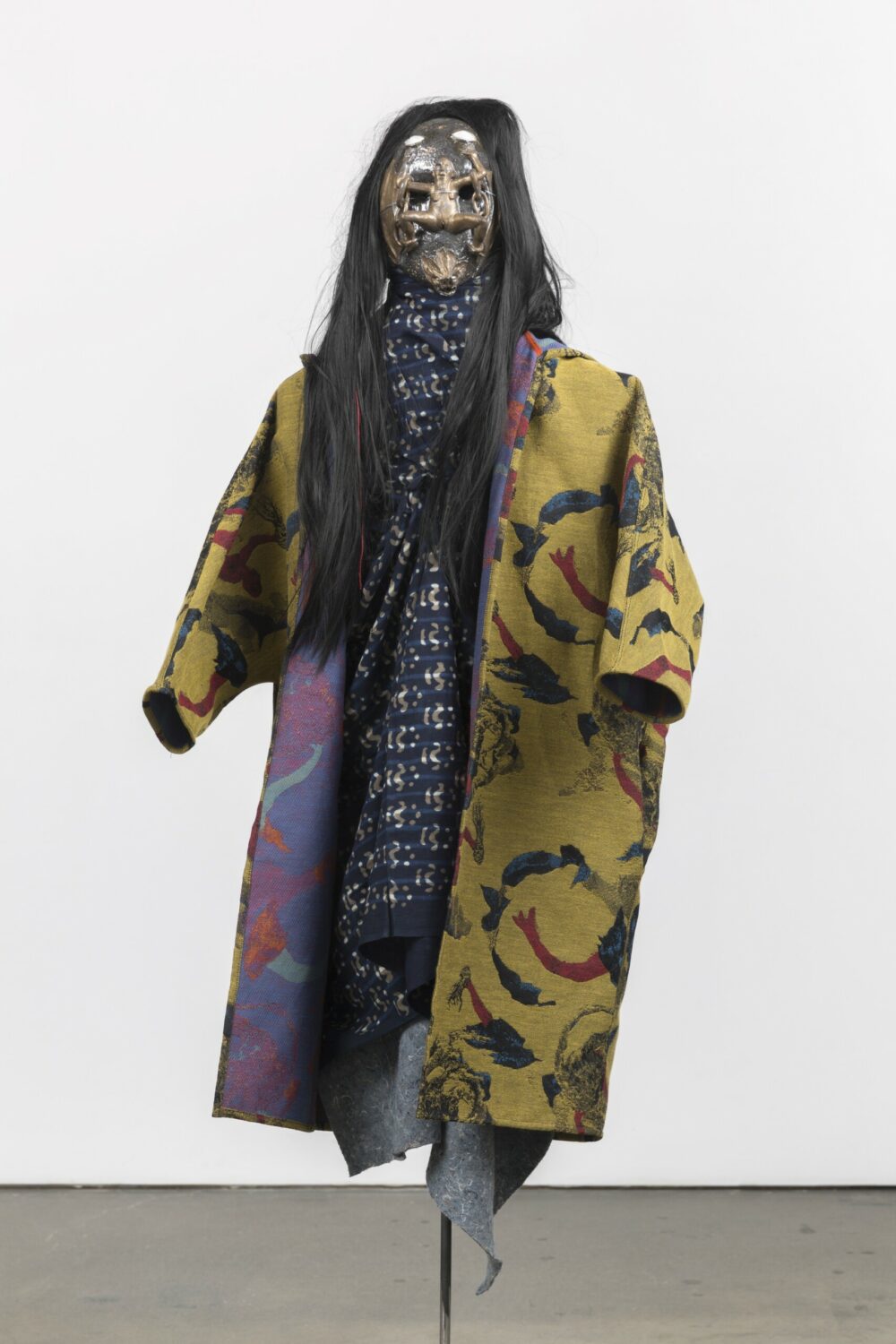 A figure or mannequin wearing a patterned yellow fabric jacket and a long patterned blue scarf, a black wig and a glazed ceramic mask