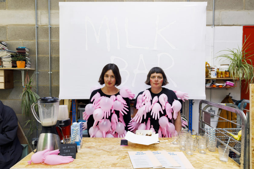 In an industrial studio setting, two women with bobbed brown hair sit in front of the camera, not smiling. They are wearing a matching outfit of a black t shirt adorned with pink plastic gloves that have been inflated. Behind them is a large sign with MILK BAR written on it. In front of them is a table with glasses, a food blender, straws, and papers.