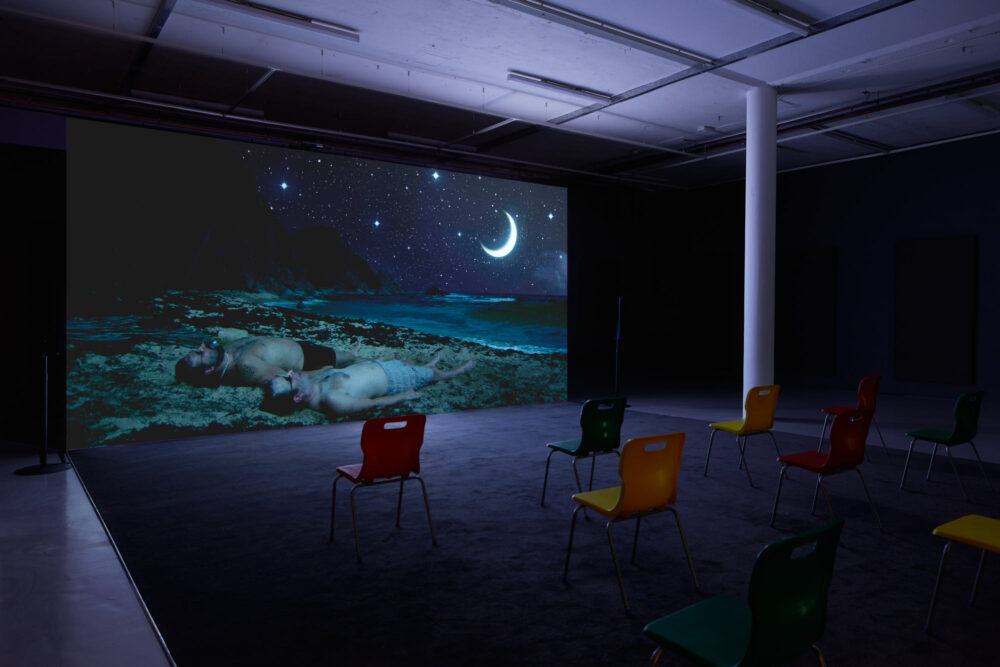 Gallery view of a projection screen showing two men wearing only shorts, lying on a beach at night, under a huge crescent moon in a starlit sky.