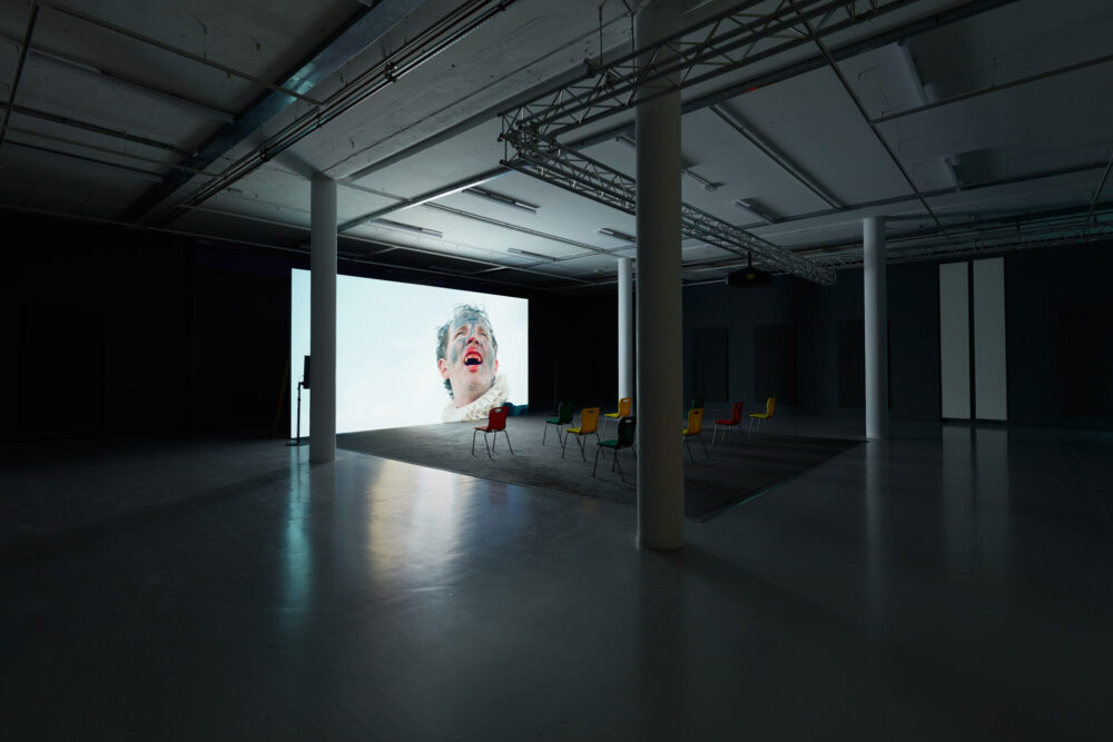 Gallery view of a projection screen showing a close up of a man, his mouth open wide in a bellow, revealing his fangs.