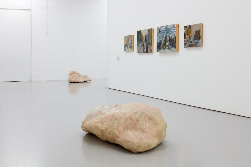 Gallery view of a large white flesh lump in the foreground and four paintings on the wall above.