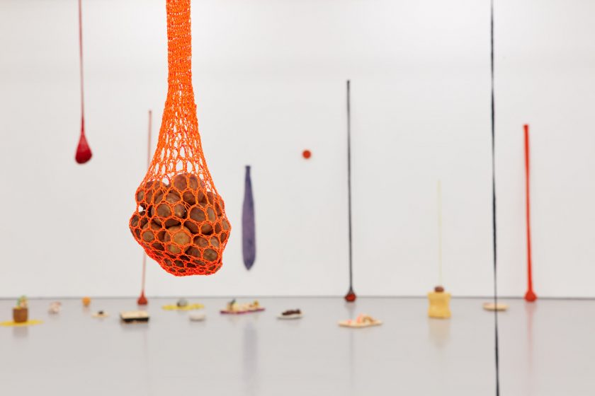 Veronica Ryan, Along a Spectrum exhibition: a white gallery space filled with hanging colourful net pouches and a variety of colourful objects sit on the floor.