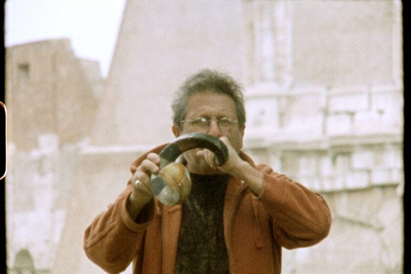 Film still featuring a man with white hair and round glasses and wearing a black tshirt, blowing into an animal horn