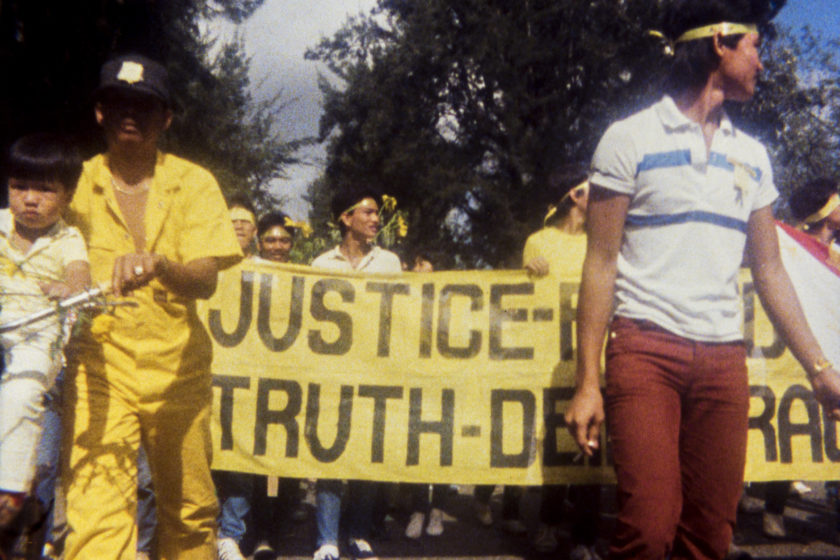 A grainy photograph of a protest. People in the background are wearing yellow headbands and holding yellow flyers. They are also displaying a large banner where the words 