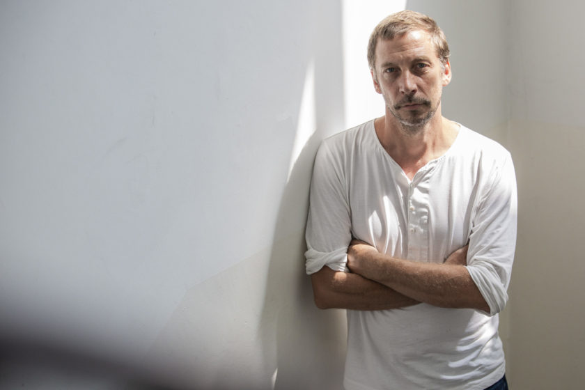 Artist Eric Baudelaire is photographed standing against a grey wall, with his arms folded across his chest and he is wearing a white long sleeved top.