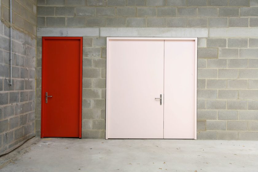 A photograph of a grey breeze block wall, featuring two closed doors - the left door is red and the right door is pink.