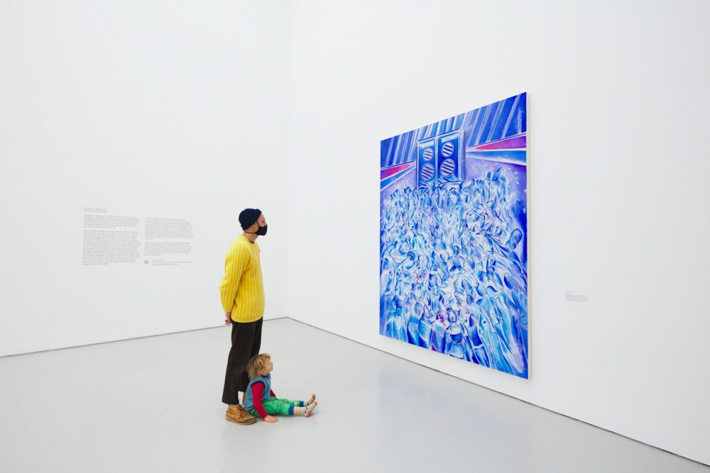 A photograph of a man and small child in a bright white gallery looking at the Denzil Forrester painting Itchin & Scratchin, a vibrant blue painting of bodies moving to music in a nightclub.