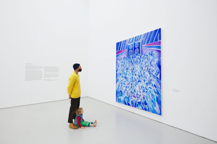 A photograph of a man and small child in a bright white gallery looking at the Denzil Forrester painting Itchin & Scratchin, a vibrant blue painting of bodies moving to music in a nightclub.