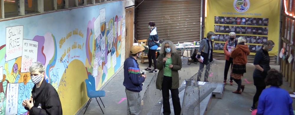 Anna Haydock-Wilson. A photograph taken from the ceiling height, of a warehouse-like space where a temporary exhibition is installed. A long colourful mural is painted on the wall on the left, and people wearing face masks are standing around the space chatting.
