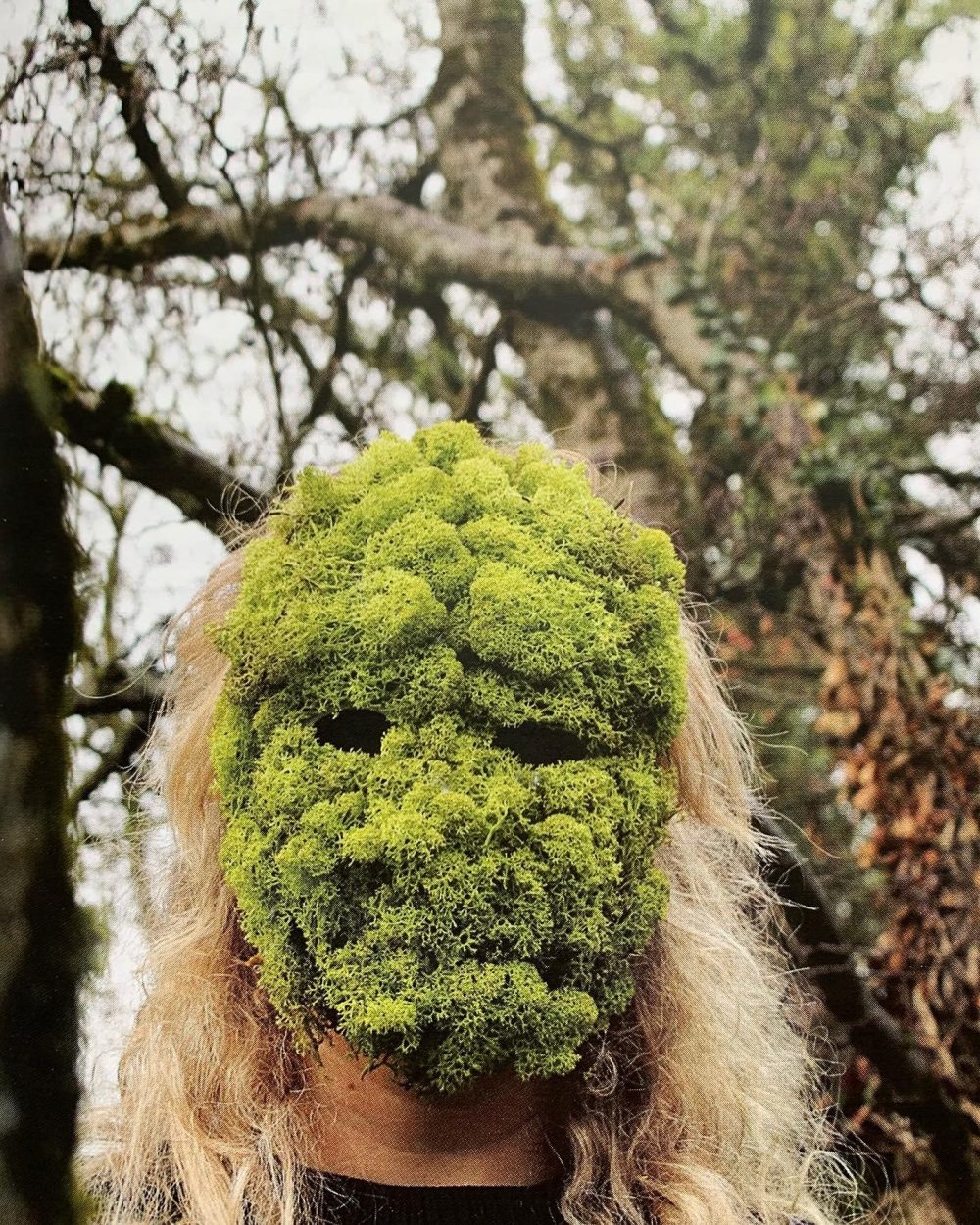 Annabel Pettigrew. A grainy colour photograph of a person with long light blonde hair, positioned in a forest setting, with a mask over their face made from bright green moss.