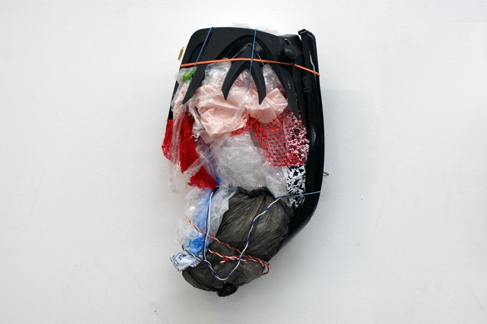 Ben Hartley. an oval-shaped sculpture comprising of pink and grey plastic bags, clear bubble wrap; blue, white, and red wire, clear tape, red netting and other unidentified objects hung on a white wall