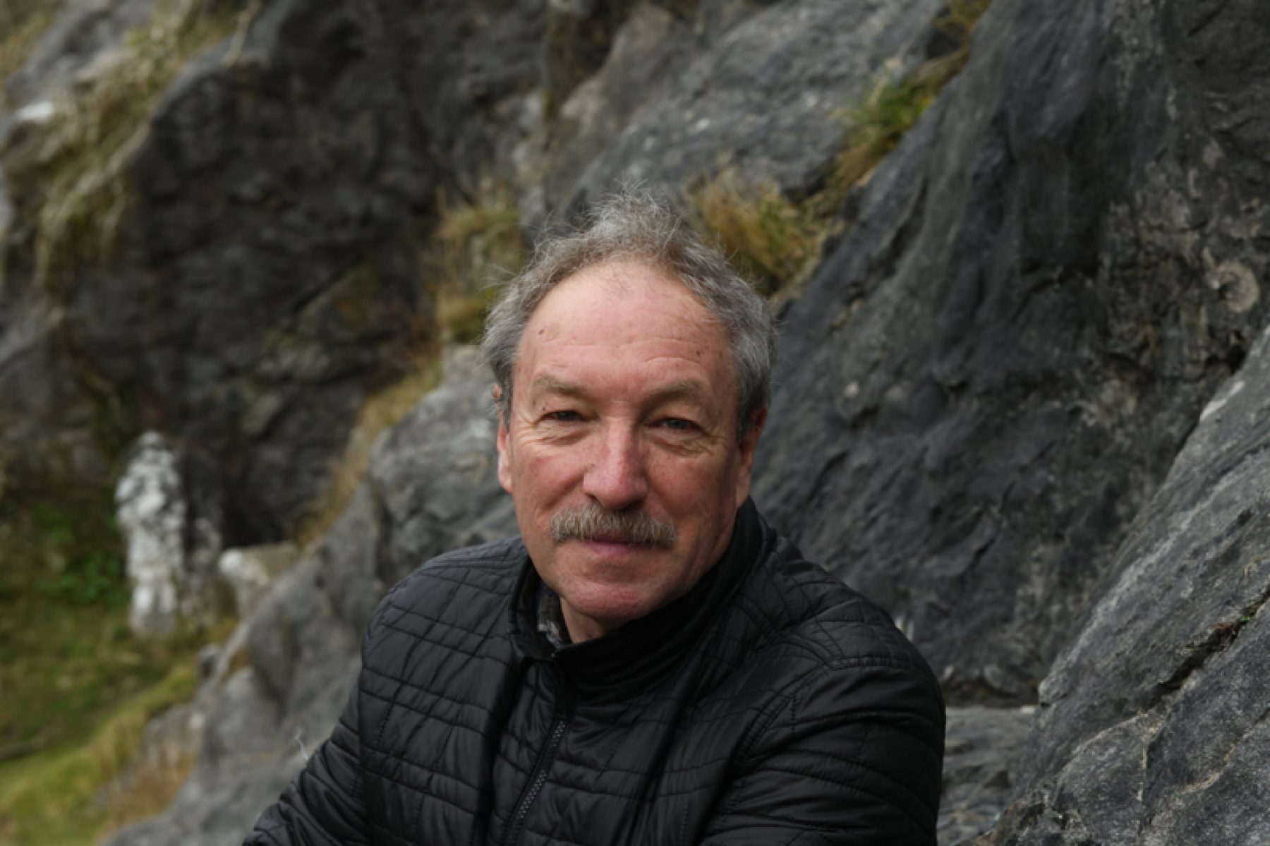 A white man with grey hair and moustache, wearing a black puffer jacket sits in front of grey rock forms with his hands in his pockets.