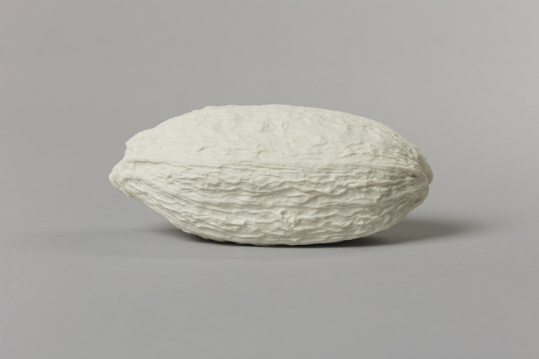 An off-white porcelain cast cocoa pod – an elongated spheroid with a crinkled and textured surface – photographedon a grey background