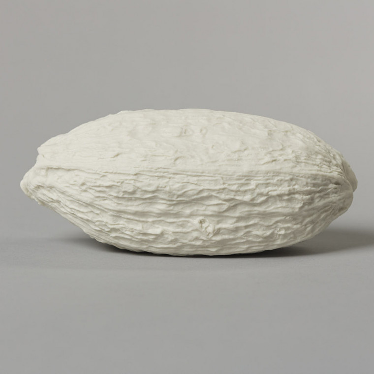 An off-white porcelain cast cocoa pod – an elongated spheroid with a crinkled and textured surface – photographedon a grey background