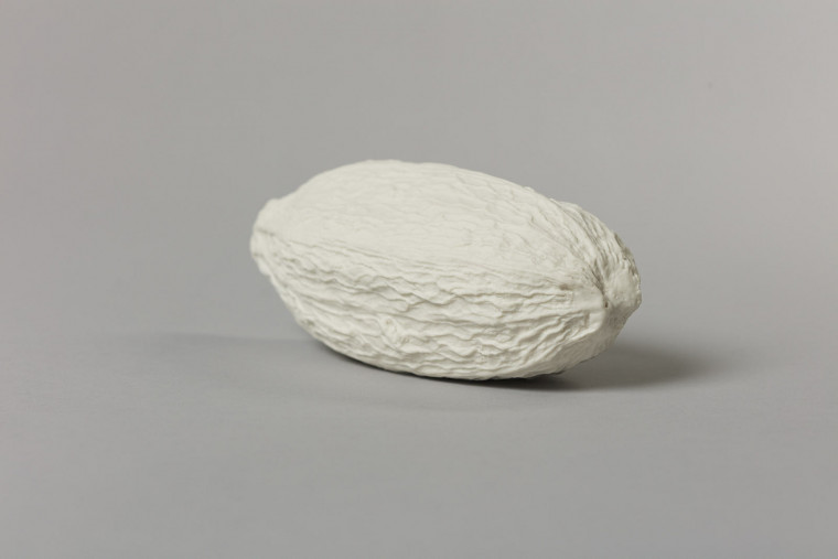 An off-white porcelain cast cocoa pod – an elongated spheroid with a crinkled and textured surface – photographedon a grey background