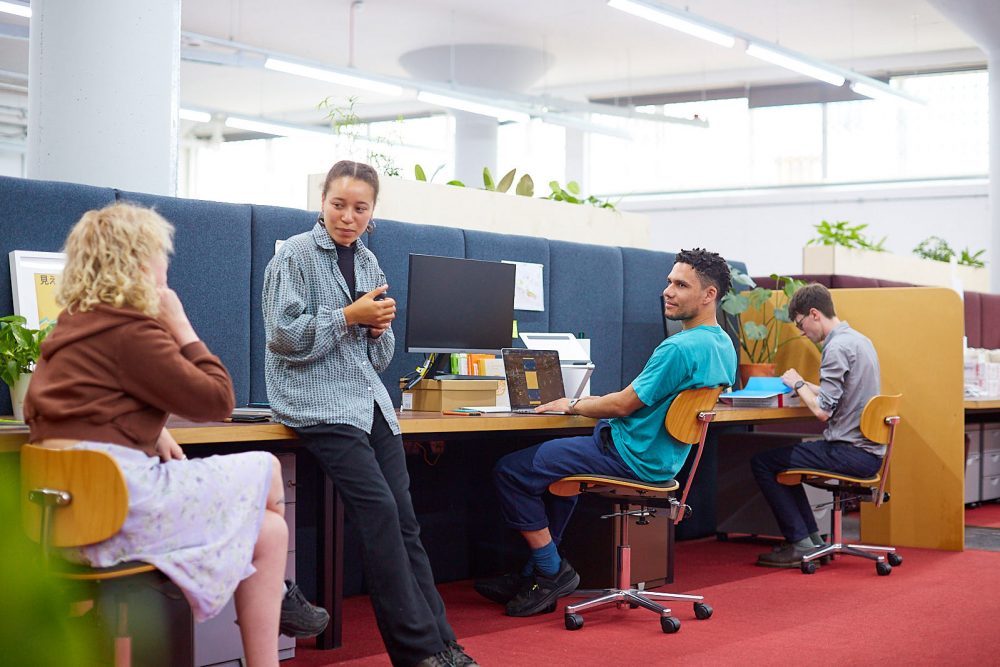 A group of people chatting in a co-working space, sitting in a row, one person standing with a cup in their hands.