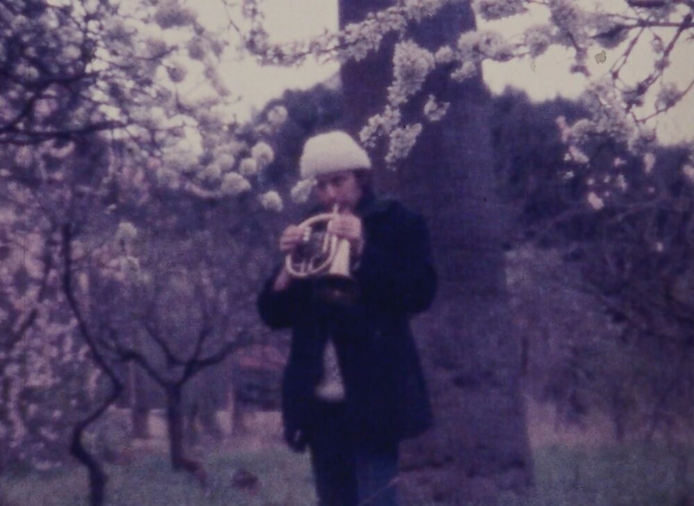 Film still of a man playing a small brass instrument in front of a blossom tree, wearing a white woolly hat