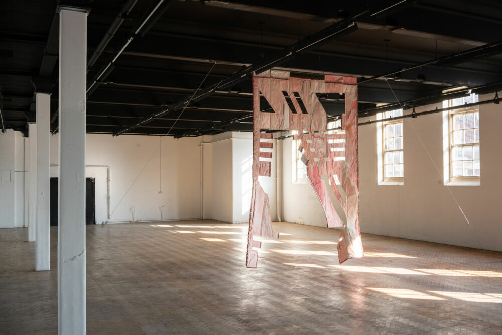 A large light pink flag with geometric cut-away panels hanging in a large empty room with wooden floor.