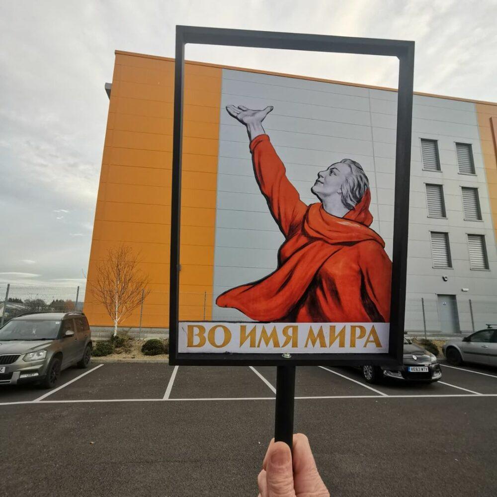 In the foreground of this image there is a stencil of a woman wearing a red headscarf and top, with her hand in the air looking upwards as if she is exclaiming. In the background there is a grey and orange warehouse building and a car park.