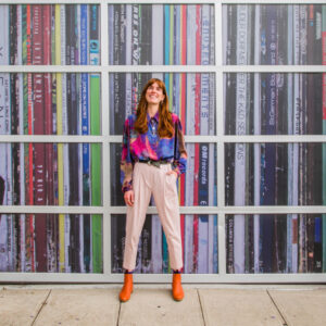 A woman smiling with long ginger hair, cream trousers and a coulourful blouse, standing in front of a colourful wall.