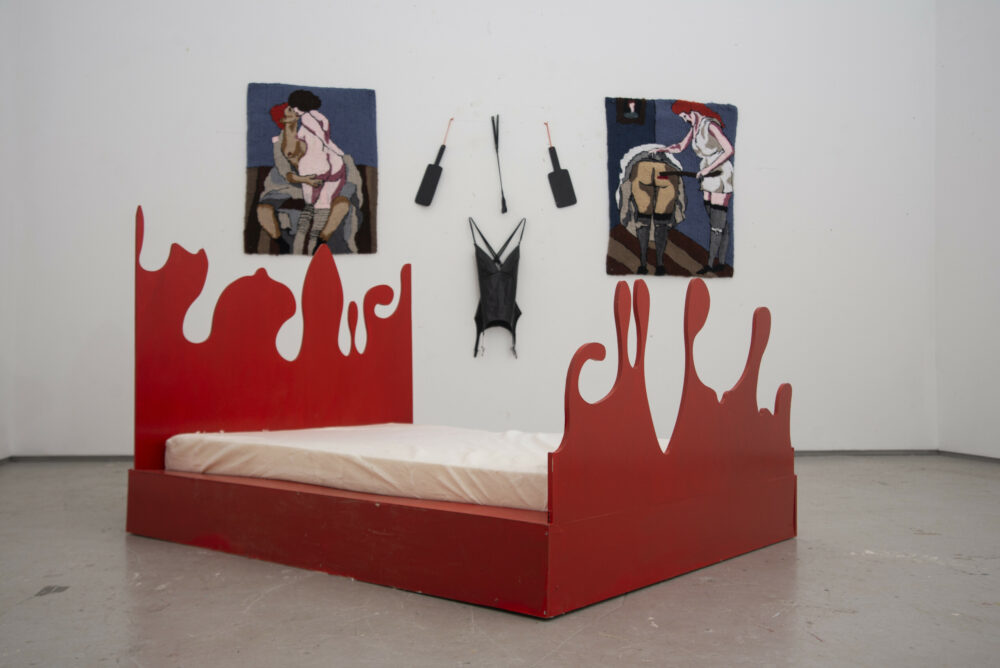 An installation with a red bed in the middle and a series of tapestries and wall hangings behind