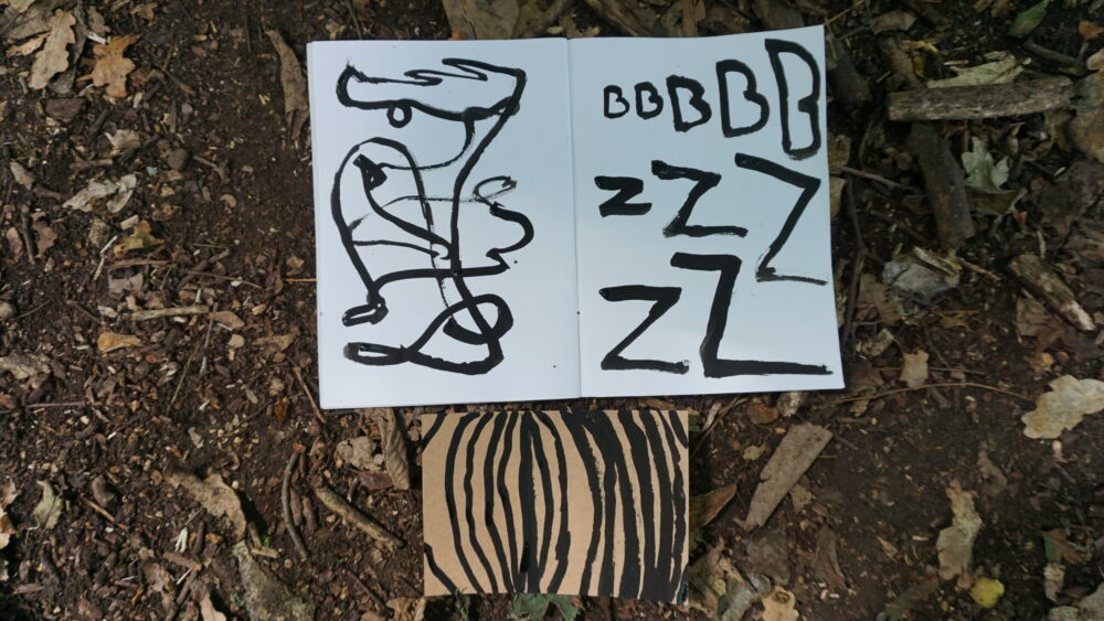 image of a woodland floor where a notebook lays, with black lines and letters painted on the pages