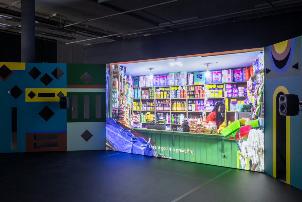 A film still of the counter of a brightly-coloured shop is presented on a large screen.