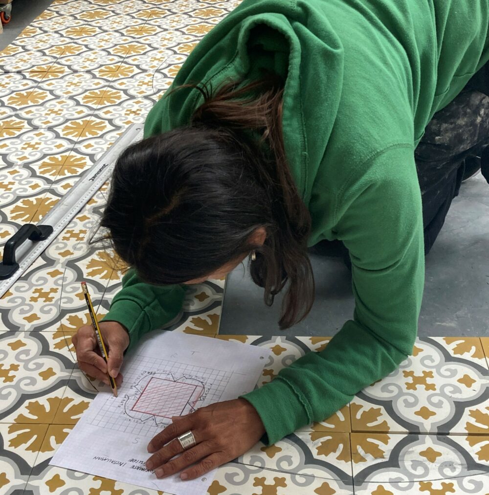 Artist Huma Mulji kneeling on a tiled floor to draw on a piece of paper.