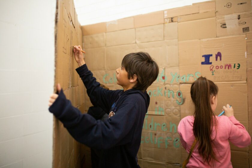 Image of two children drawing and painting on a wall made of cardboard.