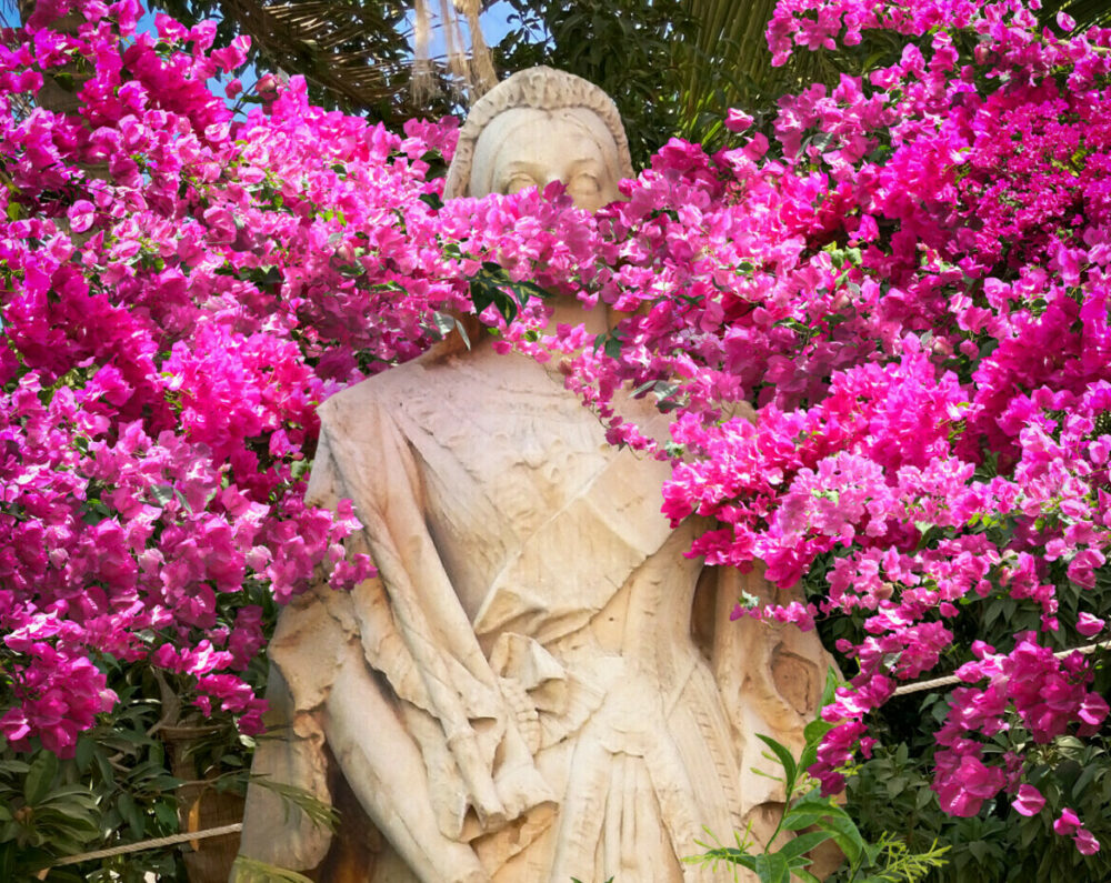 A historical statue of a female figure outdoors, obscured by plants and pink flowers.