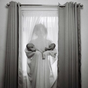 person draped in chiffon curtain standing against window holding two babies in each arm.