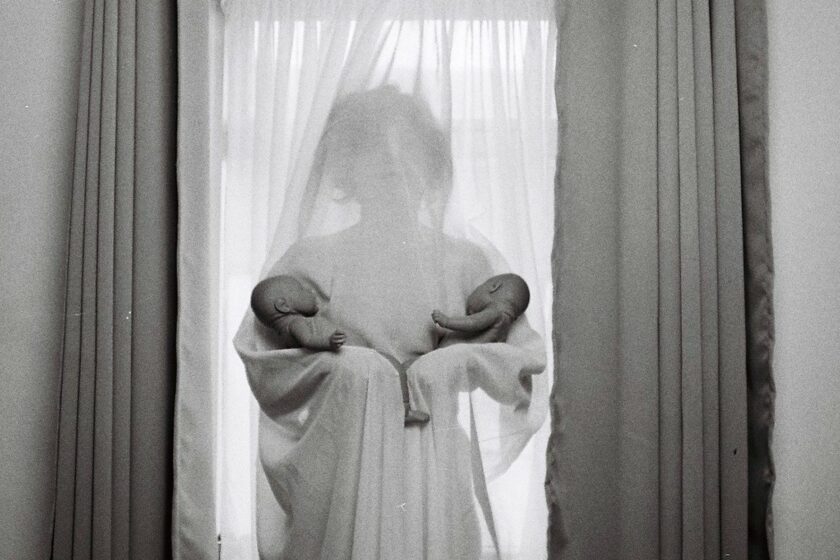person draped in chiffon curtain standing against window holding two babies in each arm.