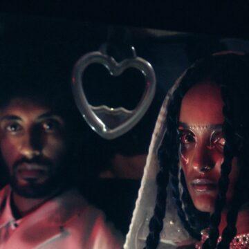 A close up of two people in dressed white decorative clothes and jewellery sitting in a car at night time in bright pink light.