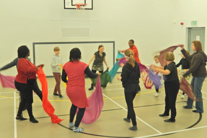 A group of adults and children stood in a circle waving different coloured scarves as part of a workshop.