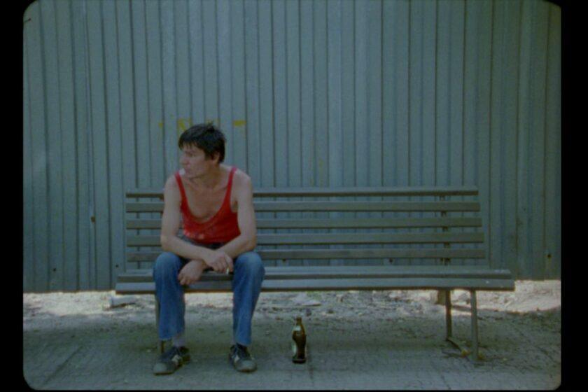 A man in a red tank top and jeans sitting on a bench against a grey wall.